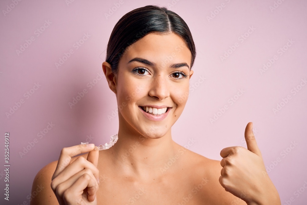 Young beautiful girl holding dental aligner over isolated pink background happy with big smile doing ok sign, thumb up with fingers, excellent sign