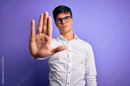Young handsome business man wearing shirt and glasses over isolated purple background doing stop sing with palm of the hand. Warning expression with negative and serious gesture on the face.