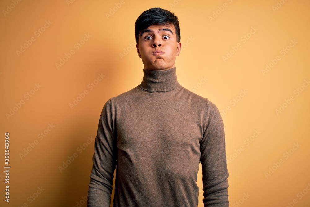 Young handsome man wearing casual turtleneck sweater over isolated yellow background puffing cheeks with funny face. Mouth inflated with air, crazy expression. Young handsome man wearing casual turtleneck sweater over isolated yellow background puffing cheeks with funny face. Mouth inflated with air, crazy expression.