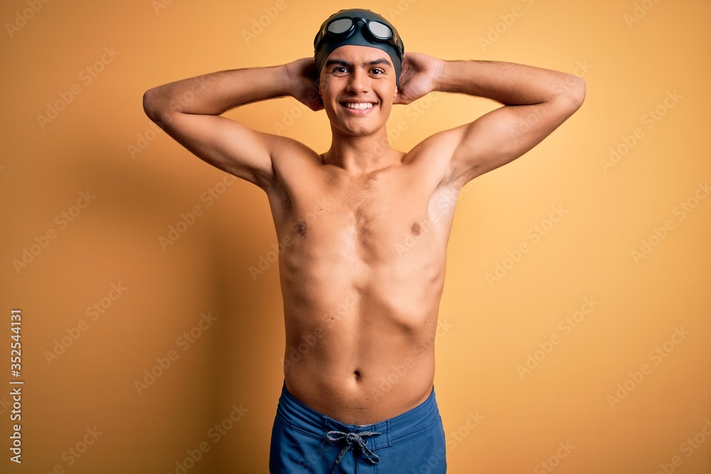 Young handsome man shirtless wearing swimsuit and swim cap over isolated yellow background relaxing and stretching, arms and hands behind head and neck smiling happy
