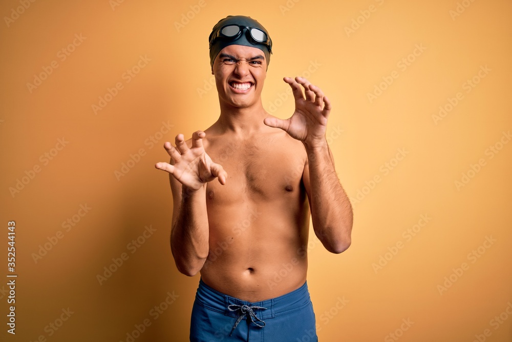 Young handsome man shirtless wearing swimsuit and swim cap over isolated yellow background smiling funny doing claw gesture as cat, aggressive and sexy expression
