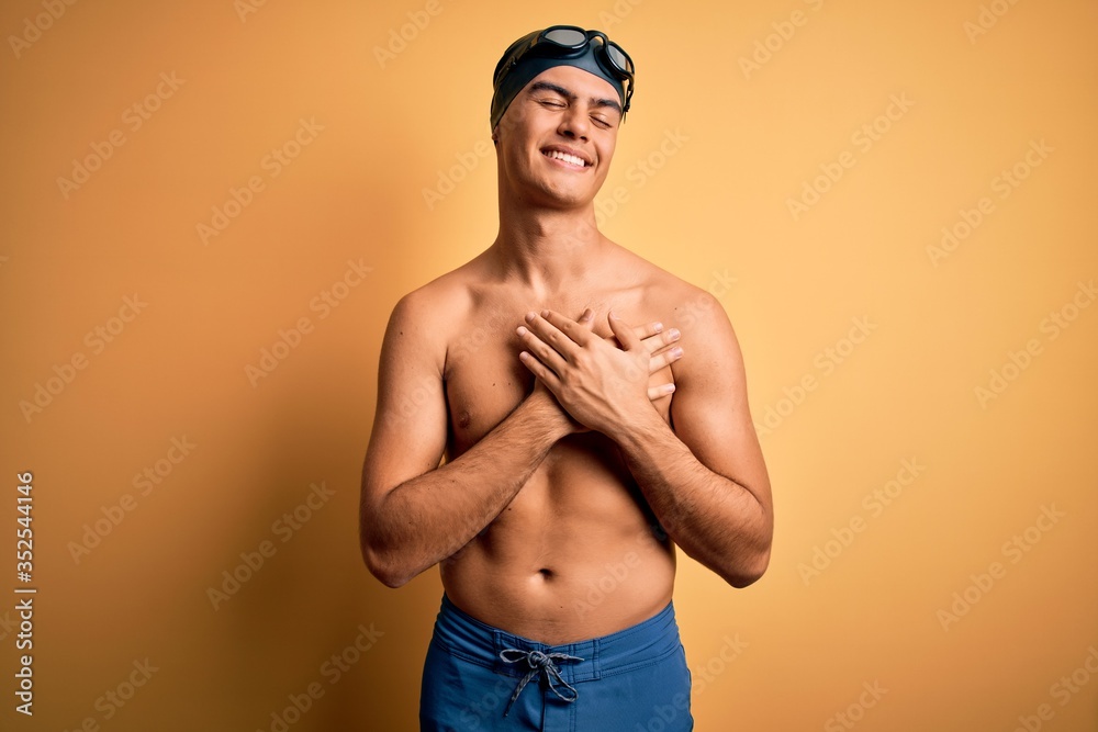 Young handsome man shirtless wearing swimsuit and swim cap over isolated yellow background smiling with hands on chest with closed eyes and grateful gesture on face. Health concept.