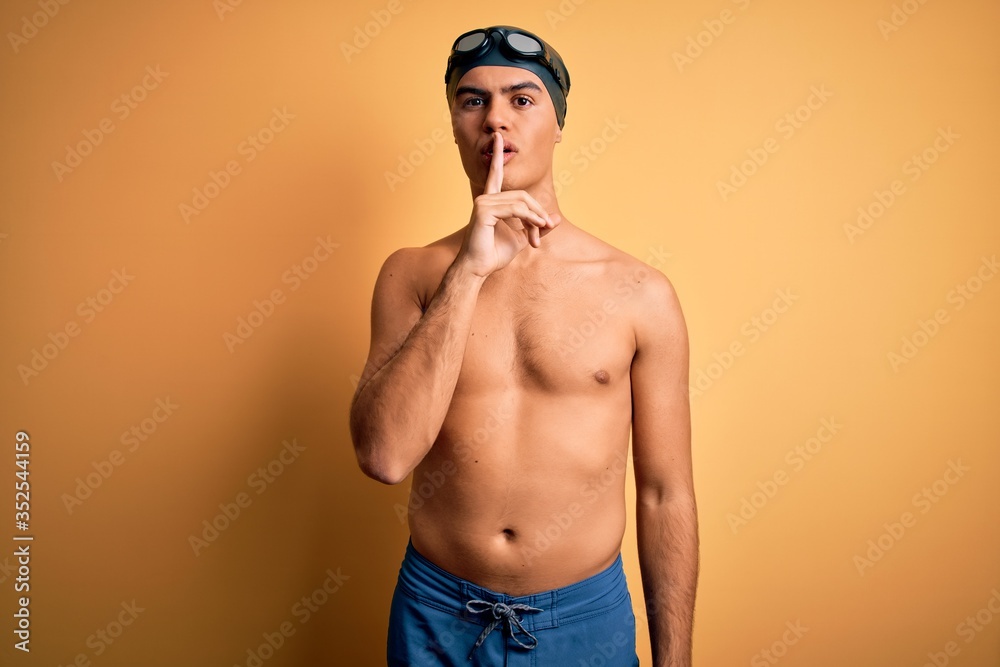 Young handsome man shirtless wearing swimsuit and swim cap over isolated yellow background asking to be quiet with finger on lips. Silence and secret concept.