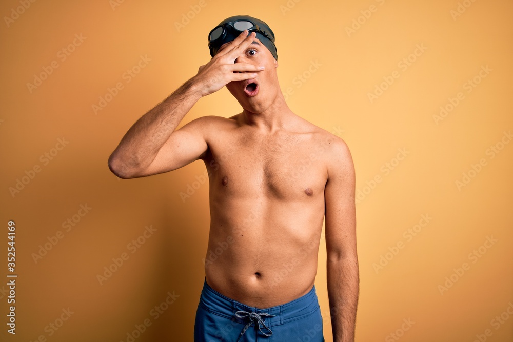 Young handsome man shirtless wearing swimsuit and swim cap over isolated yellow background peeking in shock covering face and eyes with hand, looking through fingers with embarrassed expression.
