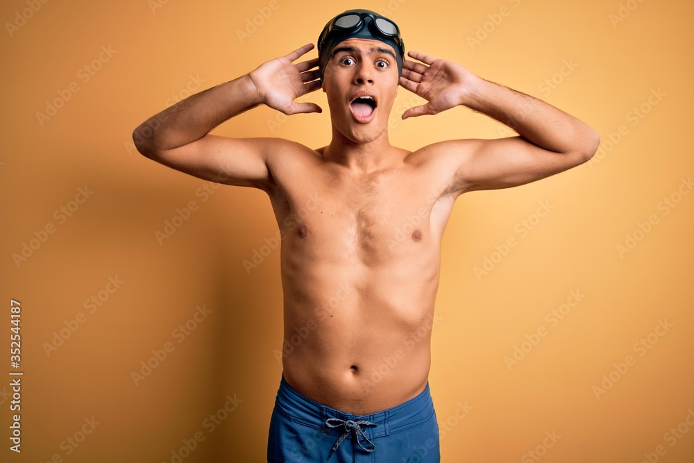 Young handsome man shirtless wearing swimsuit and swim cap over isolated yellow background Crazy and scared with hands on head, afraid and surprised of shock with open mouth