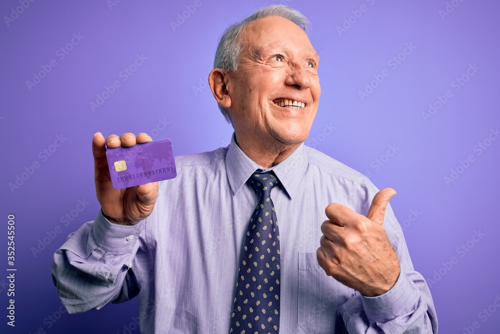 Senior grey haired business man holding credit card over purple background pointing and showing with thumb up to the side with happy face smiling Senior grey haired business man holding credit card over purple background pointing and showing with thumb up to the side with happy face smiling