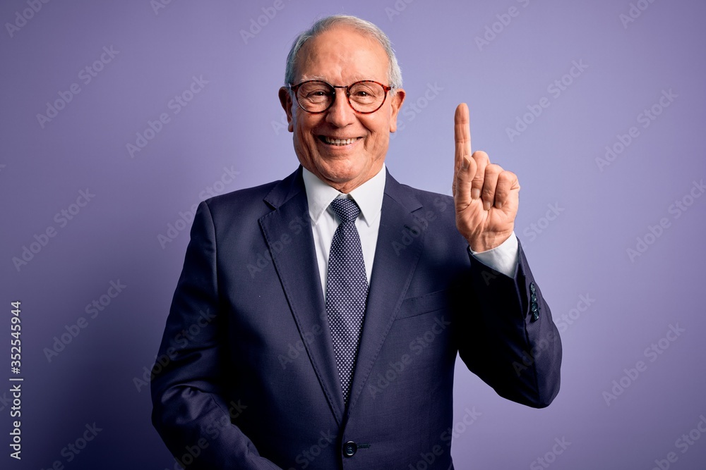 Grey haired senior business man wearing glasses and elegant suit and tie over purple background showing and pointing up with finger number one while smiling confident and happy. Grey haired senior business man wearing glasses and elegant suit and tie over purple background showing and pointing up with finger number one while smiling confident and happy.