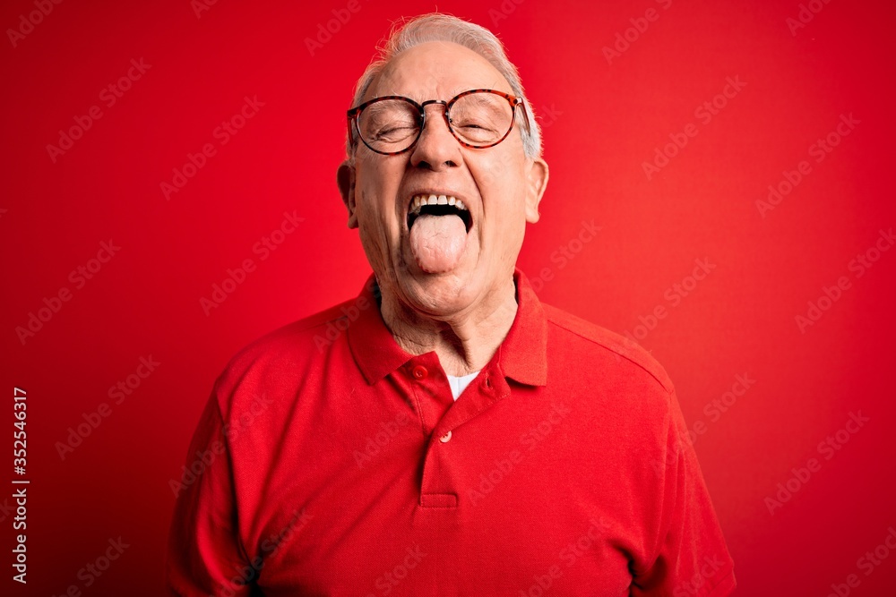 Obraz premium Grey haired senior man wearing glasses and casual t-shirt over red background sticking tongue out happy with funny expression. Emotion concept.