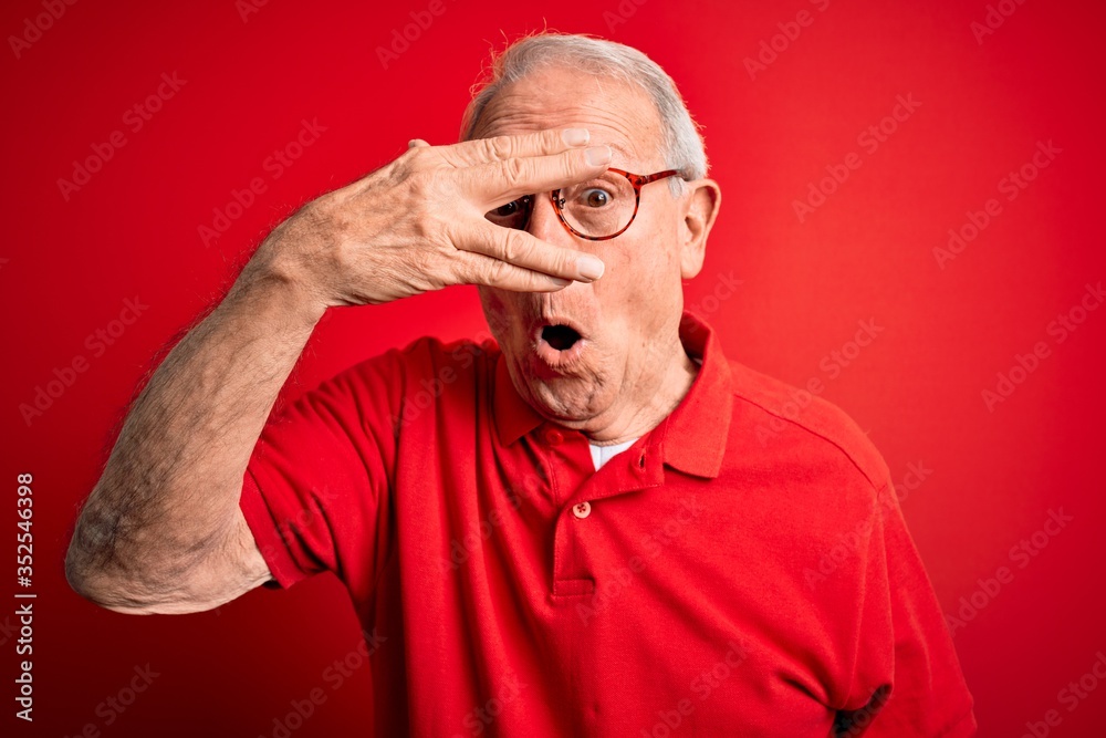 Obraz premium Grey haired senior man wearing glasses and casual t-shirt over red background peeking in shock covering face and eyes with hand, looking through fingers with embarrassed expression.
