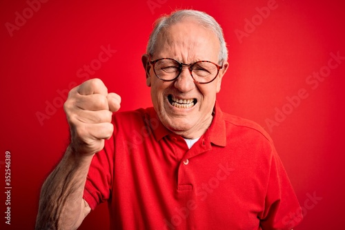 Grey haired senior man wearing glasses and casual t-shirt over red background angry and mad raising fist frustrated and furious while shouting with anger. Rage and aggressive concept.