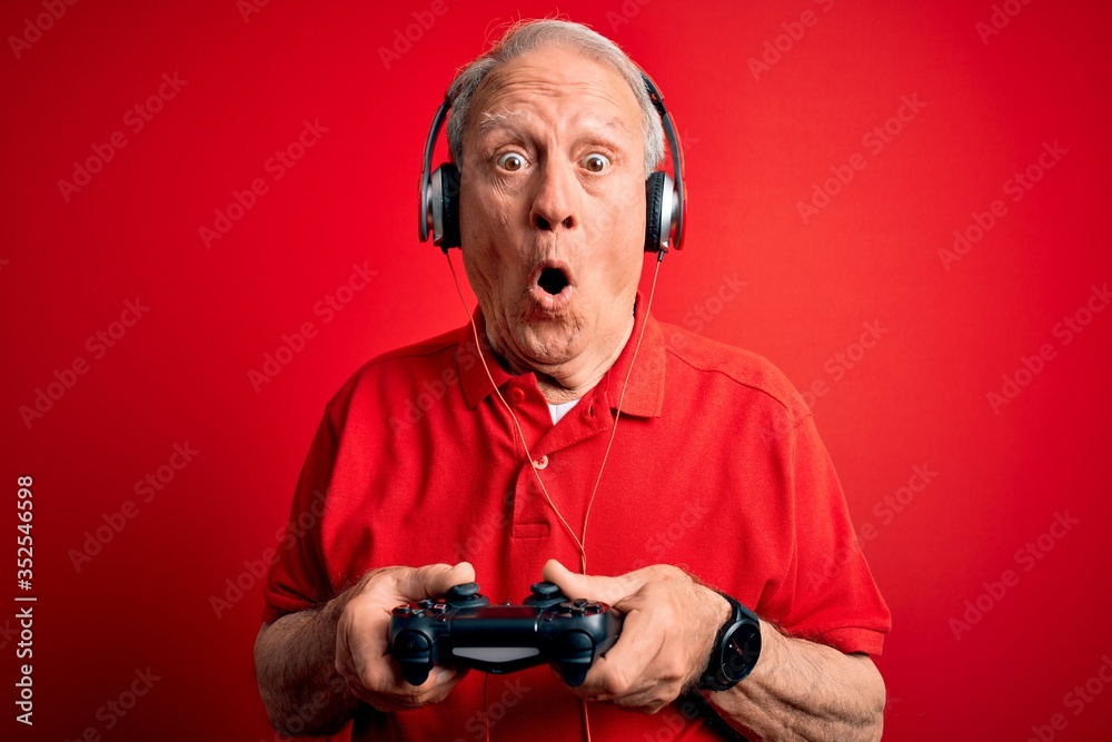 Senior grey haired gamer man playing video games using gamepad joystick over red background scared in shock with a surprise face, afraid and excited with fear expression Senior grey haired gamer man playing video games using gamepad joystick over red background scared in shock with a surprise face, afraid and excited with fear expression