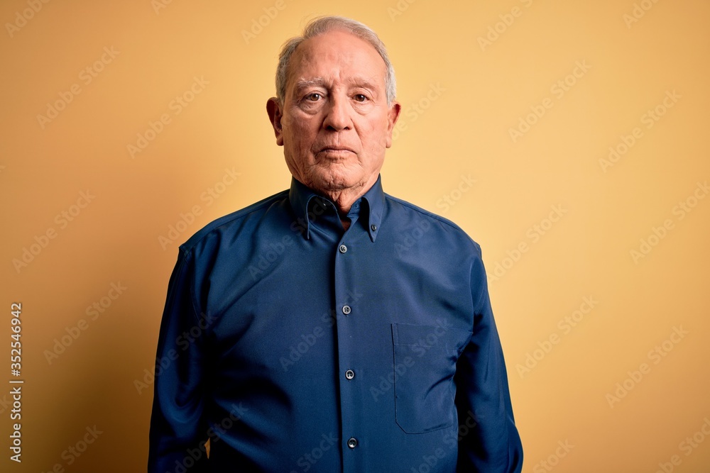 Grey haired senior man wearing casual blue shirt standing over yellow background Relaxed with serious expression on face. Simple and natural looking at the camera.