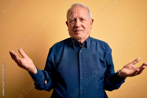 Grey haired senior man wearing casual blue shirt standing over yellow background clueless and confused with open arms, no idea concept.