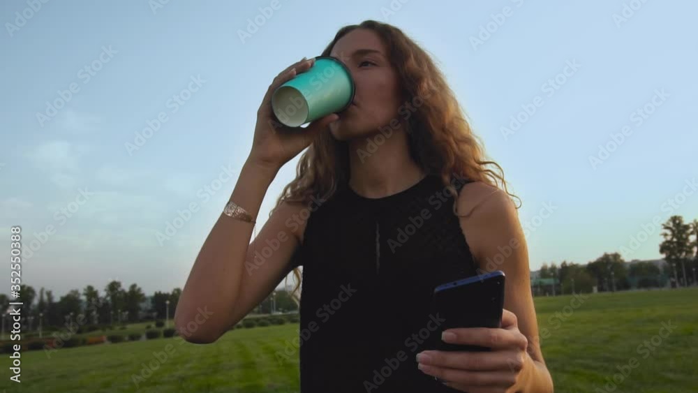 Serious curly long haired girl drinks coffee and looks at smartphone at sunset in park outdoors. Woman watching news feed, she places order in online store and makes purchase payment. Remote Shoping Serious curly long haired girl drinks coffee and looks at smartphone at sunset in park outdoors. Woman watching news feed, she places order in online store and makes purchase payment. Remote Shoping