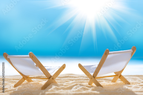 Deck chairs on sandy beach with blurry blue ocean and sun beams on sky. Social distancing or COVID-19 protection at summer holidays. Summer background. Soft focus