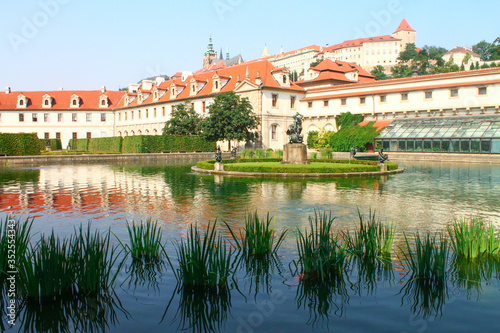 View of reflection in a lake in Wallenstein Garden, its statues and fountain, red tiled roof against vibrant blue sky and white clouds in daytime. Prague, Chezh Republic.