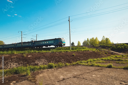 Pleasant summer landscape: old blue sky and clouds, green grass and trees, railway path road, old rusty industrial metal construction and a train. A bright scenery view and spirit of adventure.