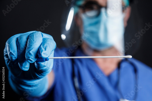 Medico  con protezioni e camice blu mostra un tampone faringeo per controllare lo stato di salute , isolato su sfondo nero photo