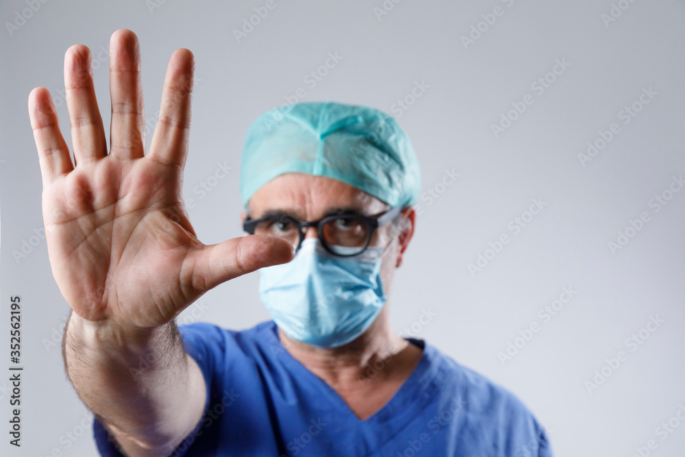 Chirurgo con protezioni e camice blu mostra la mano in segno di stop, isolato su sfondo bianco Chirurgo con protezioni e camice blu mostra la mano in segno di stop, isolato su sfondo bianco