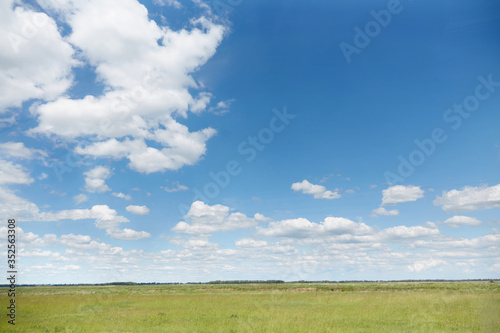 Green Sunny meadow with grass and blue summer sky with white air clouds. Beautiful summer day in the village, horizontal photo place for text