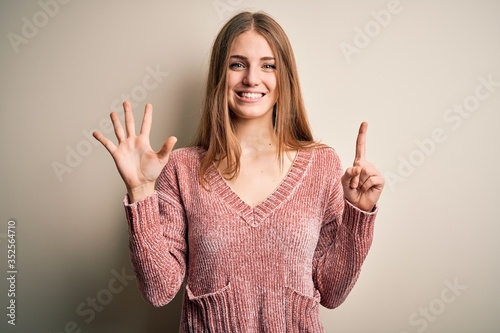 Young beautiful redhead woman wearing pink casual sweater over isolated white background showing and pointing up with fingers number six while smiling confident and happy.
