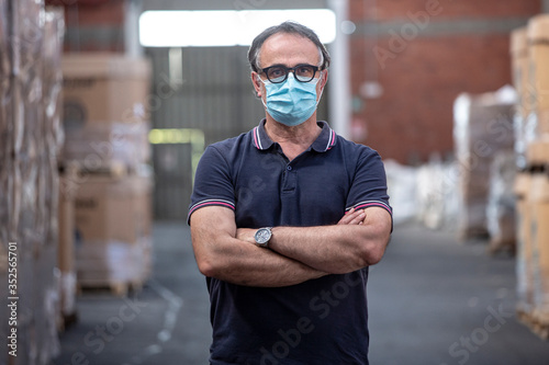 Uomo con occhiali neri e mascherina protettiva incrocia le braccia nel magazzino in cui lavora photo