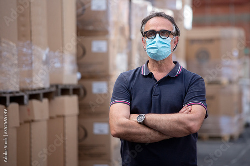 Uomo con occhiali neri e mascherina protettiva incrocia le braccia nel magazzino in cui lavora photo