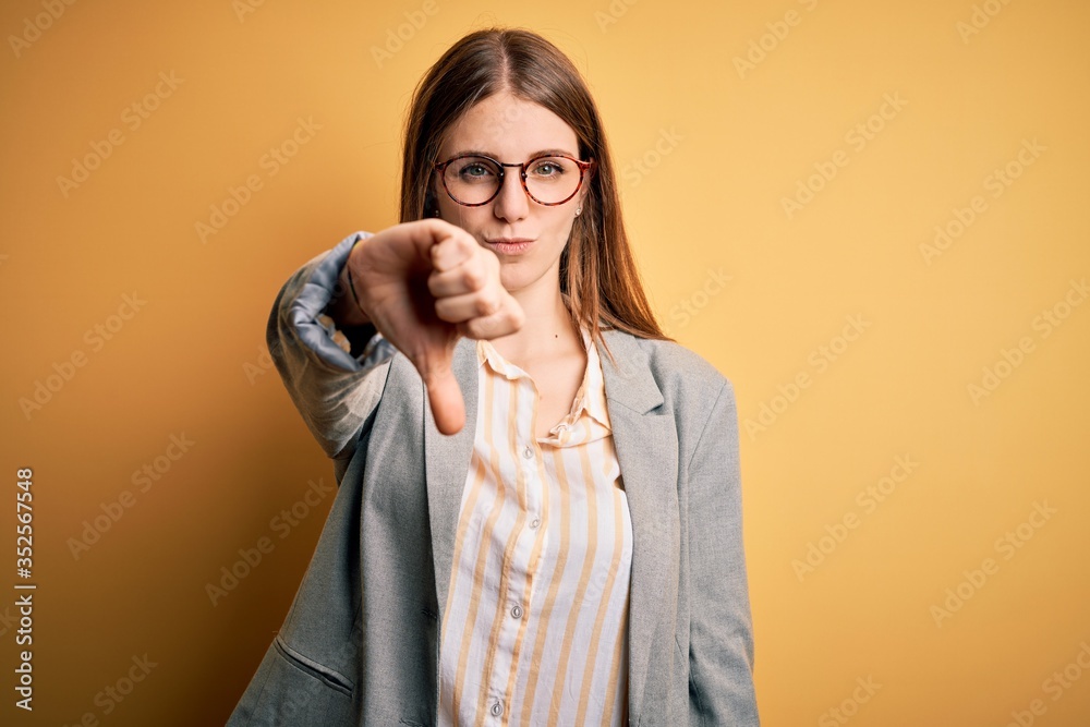 Young beautiful redhead woman wearing jacket and glasses over isolated yellow background looking unhappy and angry showing rejection and negative with thumbs down gesture. Bad expression. Young beautiful redhead woman wearing jacket and glasses over isolated yellow background looking unhappy and angry showing rejection and negative with thumbs down gesture. Bad expression.
