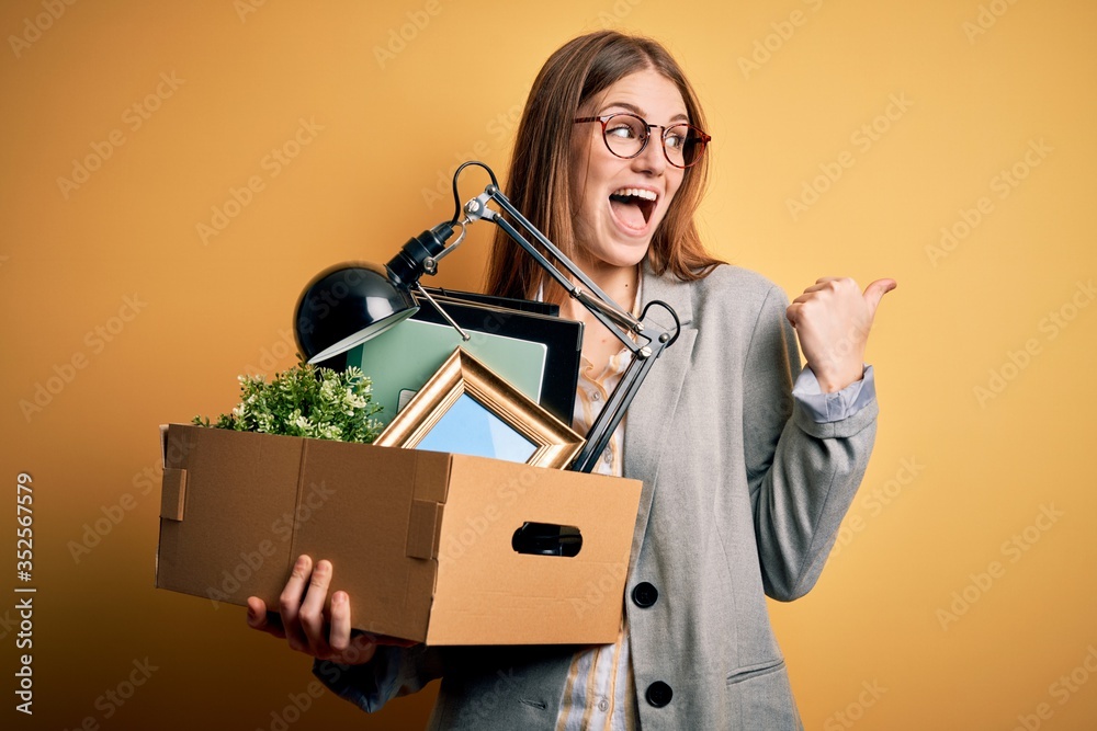 Young beautiful redhead businesswoman fired holding carboard box over yellow background pointing and showing with thumb up to the side with happy face smiling Young beautiful redhead businesswoman fired holding carboard box over yellow background pointing and showing with thumb up to the side with happy face smiling