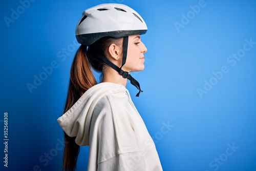 Young beautiful redhead cyclist woman wearing bike helmet over isolated blue background looking to side, relax profile pose with natural face with confident smile.