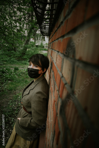 Young beautiful brunette girl in a black medical mask on a background of trees and a red brick wall. Protection health from the coronovirus. Pandemic Coronavirus Quarantine. Covid 19.