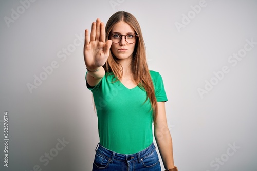 Young beautiful redhead woman wearing casual green t-shirt and glasses over white background doing stop sing with palm of the hand. Warning expression with negative and serious gesture on the face.