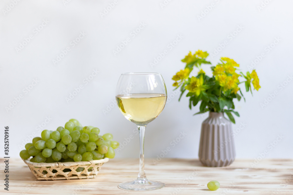 Glass of white wine, green grape in basket, yellow flowers bouquet in vasw on wooden table