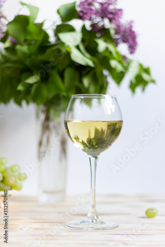 White wine in glass reflecting lilac bouquet and grape on wooden background