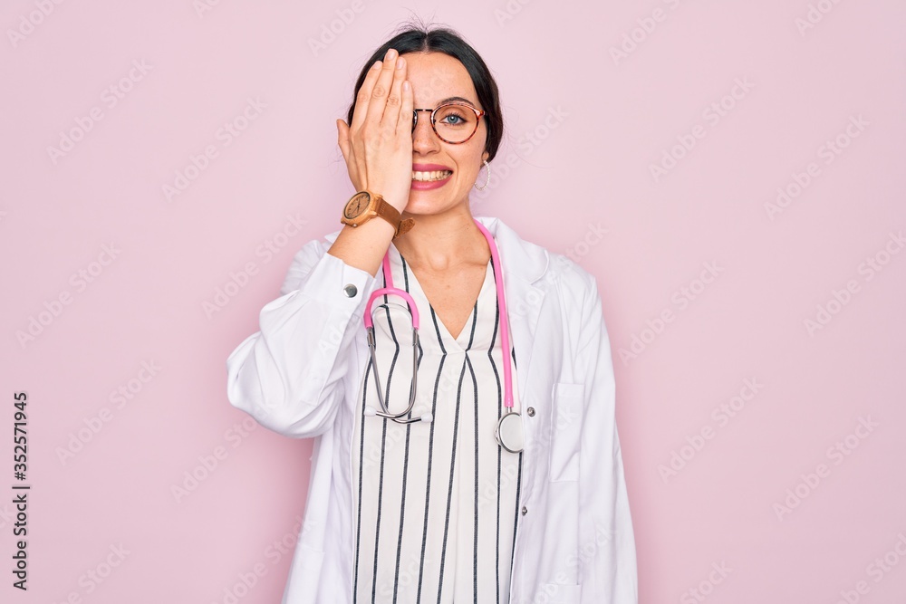 Beautiful doctor woman with blue eyes wearing coat and stethoscope over pink background covering one eye with hand, confident smile on face and surprise emotion. Beautiful doctor woman with blue eyes wearing coat and stethoscope over pink background covering one eye with hand, confident smile on face and surprise emotion.