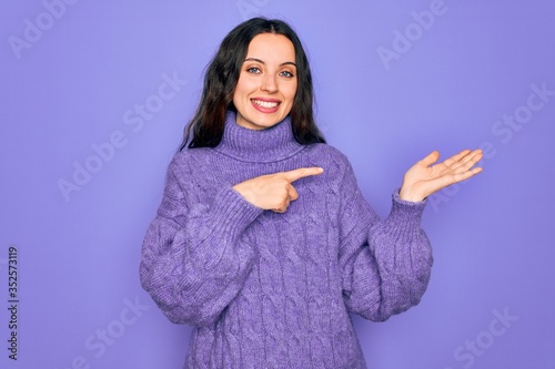 Young beautiful woman wearing casual turtleneck sweater standing over purple background amazed and smiling to the camera while presenting with hand and pointing with finger. © Krakenimages.com