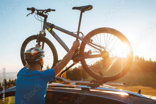 Young Man dresses modern cycling clothes and protective helmet installing his mountain bike on the car roof with sunset backlight. Active sporty people concept image.