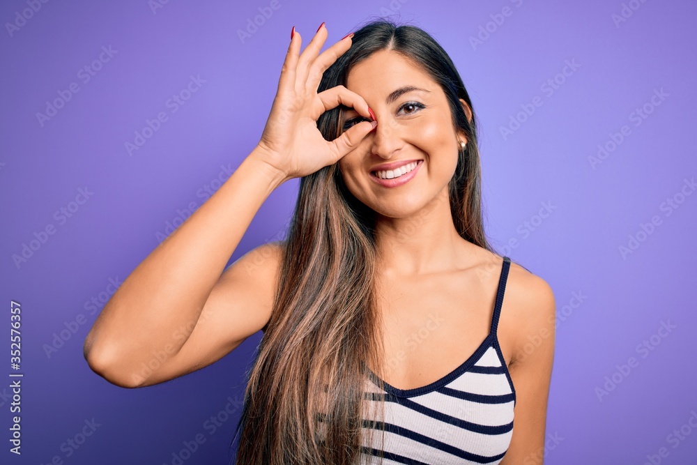 Young beautiful woman wearing casual summer striped top over purple isolated background doing ok gesture with hand smiling, eye looking through fingers with happy face. Young beautiful woman wearing casual summer striped top over purple isolated background doing ok gesture with hand smiling, eye looking through fingers with happy face.