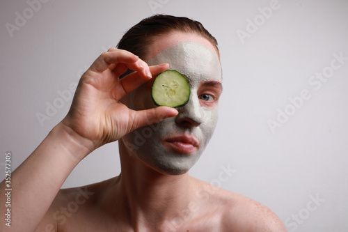 Young woman with a face mask on holding a cucumber slices in front of one eye. Beauty and skincare concept.