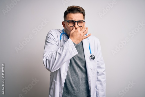 Young doctor man with blue eyes wearing medical coat and stethoscope over isolated background smelling something stinky and disgusting, intolerable smell, holding breath with fingers on nose © Krakenimages.com