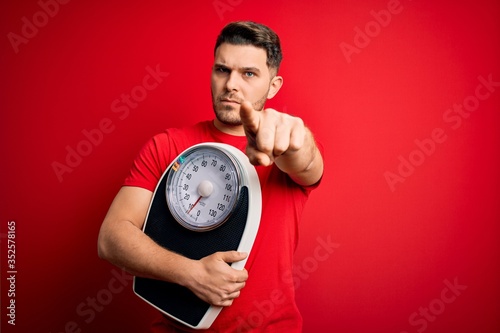 Young fitness man with blue eyes holding scale dieting for healthy weight over red background pointing with finger to the camera and to you, hand sign, positive and confident gesture from the front
