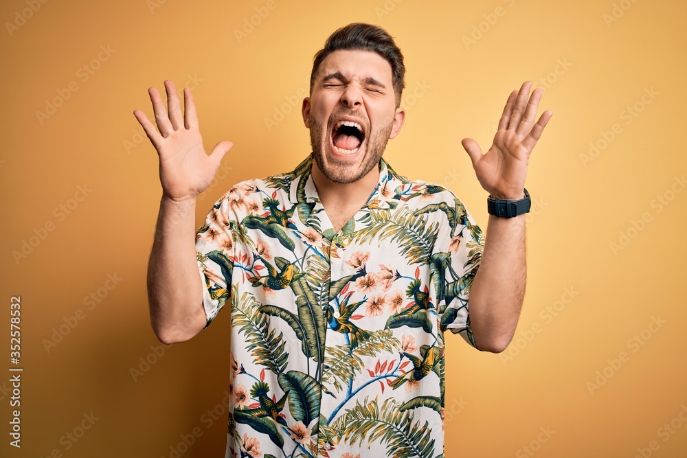Young man with blue eyes on vacation wearing floral summer shirt over yellow background celebrating mad and crazy for success with arms raised and closed eyes screaming excited. Winner concept Young man with blue eyes on vacation wearing floral summer shirt over yellow background celebrating mad and crazy for success with arms raised and closed eyes screaming excited. Winner concept