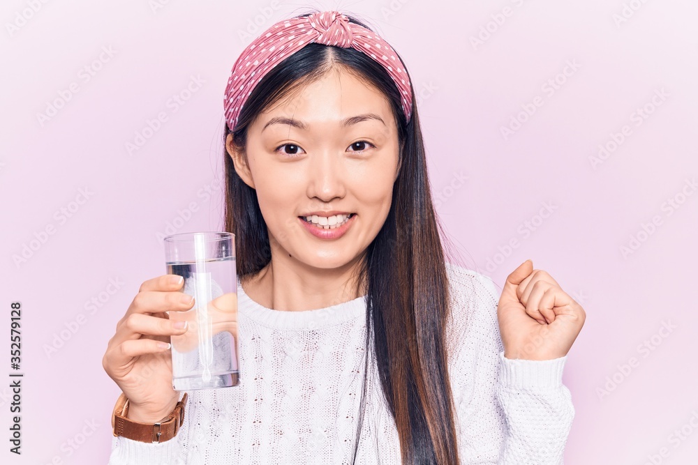 Young beautiful chinese woman drinking glass of water screaming proud, celebrating victory and success very excited with raised arm Young beautiful chinese woman drinking glass of water screaming proud, celebrating victory and success very excited with raised arm