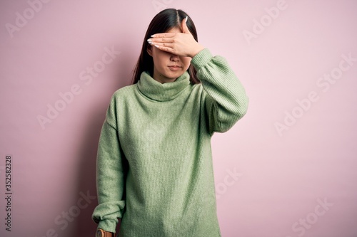 Young beautiful asian woman wearing green winter sweater over pink solated background covering eyes with hand, looking serious and sad. Sightless, hiding and rejection concept © Krakenimages.com