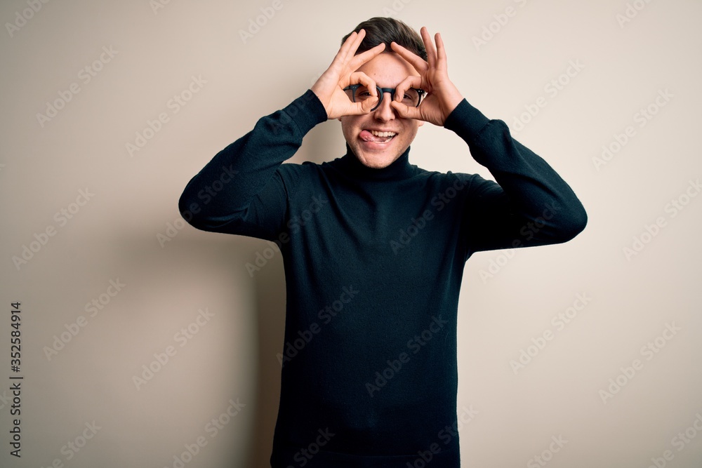Young handsome caucasian man wearing glasses and casual sweater over isolated background doing ok gesture like binoculars sticking tongue out, eyes looking through fingers. Crazy expression. Young handsome caucasian man wearing glasses and casual sweater over isolated background doing ok gesture like binoculars sticking tongue out, eyes looking through fingers. Crazy expression.