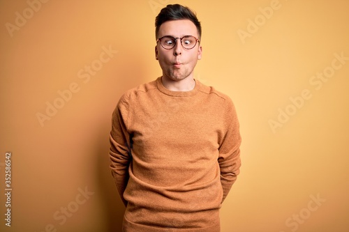 Young handsome caucasian man wearing glasses and casual winter sweater over yellow background making fish face with lips, crazy and comical gesture. Funny expression.
