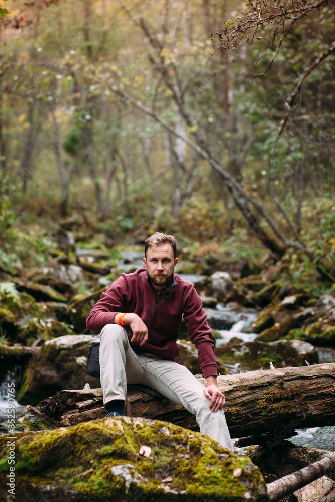 bearded young white man in maroon hoody and milky jeans sitting on the old fallen mossy tree lying abroad the river in forest bearded young white man in maroon hoody and milky jeans sitting on the old fallen mossy tree lying abroad the river in forest