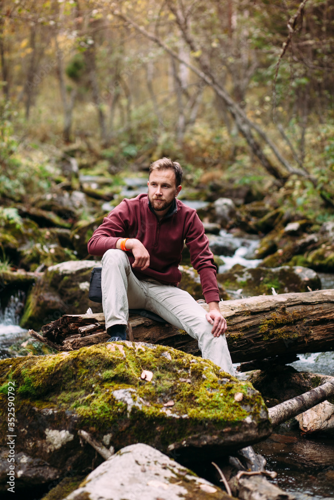 bearded young white man in maroon hoody and milky jeans sitting on the old fallen mossy tree lying abroad the river in forest
