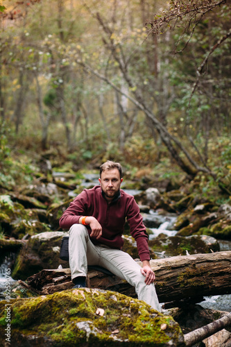 bearded young white man in maroon hoody and milky jeans sitting on the old fallen mossy tree lying abroad the river in forest