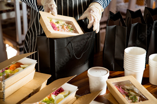 Restaurant worker wearing protective mask and gloves packing food boxed take away. Food delivery services and Online contactless food shopping.